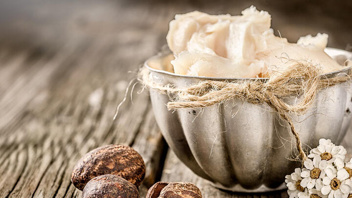 A beautiful textured bowl on an old wooden table, brimming with shea butter, with a shea seed on the left and some wildflowers on the right of the bowl
